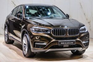 A sleek, metallic brown BMW X6 xDrive40d is parked indoors. Its front view showcases a distinctive kidney grille, LED headlights, and a chrome-accented bumper. Discover it at affordable rates through BMW Rental Las Vegas by RentaCar24.org. The car rests on large alloy wheels against a light wooden floor and a creamy marble-textured wall background.