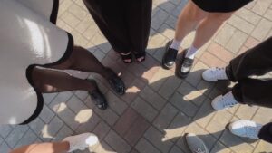 A group of people stands in a circle on a sunlit paved ground. Only their feet and legs are visible, showcasing diverse and fashionable footwear, including black heels, loafers, and white sneakers. The ground consists of gray and brown bricks forming a pattern.