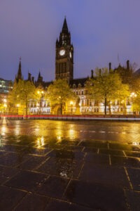 Evening view of a historic building with a tall clock tower in Manchester, illuminated by warm lights against a deep blue sky. Wet pavement reflects the glow, and trees with sparse foliage line the front. This tranquil urban scene is one of the must-see traveler experiences in the city.